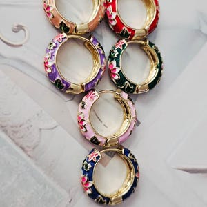 May include: A selection of enamel hoop earrings in various colours. Each earring has a gold-tone base and is decorated with floral patterns in shades of pink, red, green, and blue. The earrings are displayed on a light-coloured surface.