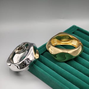 Puede incluir: Dos brazaletes gruesos y geométricos. Uno es plateado, el otro dorado. Ambos tienen un cierre de bisagra. Los brazaletes se exhiben sobre una superficie de terciopelo verde oscuro, creando un contraste de colores y texturas. El diseño es moderno y audaz.