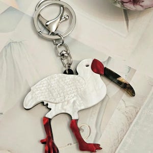 May include: A white bird keychain with red legs and a red head. The bird has a black and brown beak and is attached to a silver keyring and clasp. The bird's body has a textured, feathered appearance.