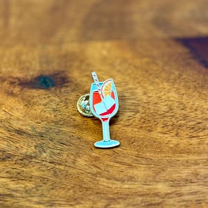 Könnte beinhalten: Ein blaues Glas mit einem roten Getränk, Eiswürfeln, einer Orangenscheibe und einem rot-weiß gestreiften Strohhalm. Das Glas steht auf einer Holzoberfläche.