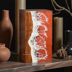 Op de afbeelding: Een rechthoekige houten kist met een opvallend ontwerp. Eén kant heeft een rijke, rood-oranje hars met een wit, schuimend patroon, dat lijkt op oceaangolven. De andere kant toont de natuurlijke nerf van het hout. De kist heeft de tekst "Vovo International".