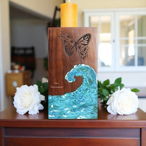 May include: A wooden candle holder with a butterfly and floral design, and a blue wave. The candle holder has a rectangular shape and a natural wood finish. A yellow candle sits on top. The text "Vina International" is visible.