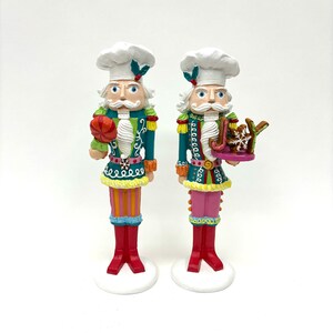 May include: Two colorful nutcracker figurines dressed as chefs. One holds a candy cane and the other holds a plate with a gingerbread cookie that says "Joy".
