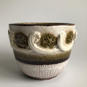 May include: A ceramic bowl featuring a cream-coloured base with a dark brown rim. The exterior is decorated with raised, swirling white scrollwork patterns and textured green floral motifs, complemented by a dark brown horizontal band.