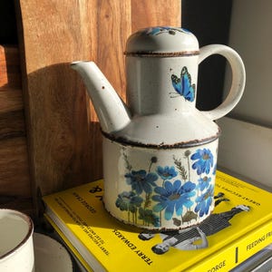 May include: A white ceramic teapot with a lid, decorated with blue butterfly and floral patterns. The teapot has a brown rim and handle. It rests on a stack of yellow cookbooks. The teapot is approximately 20 cm tall.