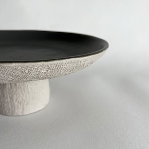 May include: A black ceramic cake stand with a textured white base. The stand has a wide, round top and a short, cylindrical base.