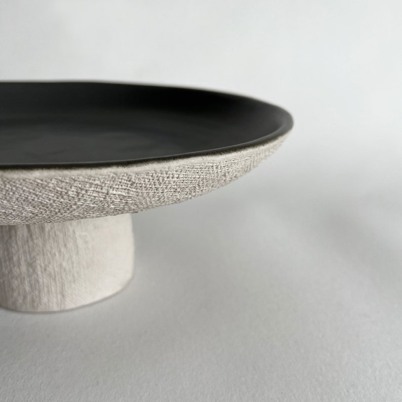 May include: A black ceramic cake stand with a textured white base. The stand has a wide, round top and a short, cylindrical base.