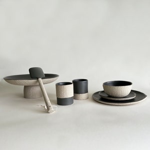 May include: A set of black and white ceramic tableware, including a serving platter, two cups, a bowl, and a spatula. The pieces have a textured surface and a minimalist design.