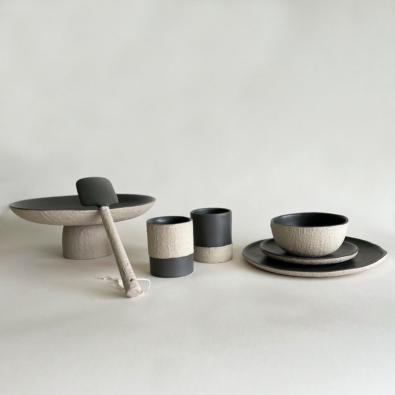 May include: A set of black and white ceramic tableware, including a serving platter, two cups, a bowl, and a spatula. The pieces have a textured surface and a minimalist design.