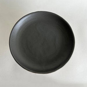 May include: A black ceramic plate with a smooth, slightly textured surface. The plate is round and has a slightly raised rim.