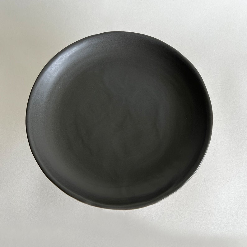 May include: A black ceramic plate with a smooth, slightly textured surface. The plate is round and has a slightly raised rim.