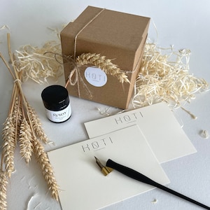 May include: A brown cardboard box with a string bow and a wheat stalk tied around it. The box has a white label with the text "HOTI LONDON" on it. There is a small black bottle of ink with a white label that says "ROUSY IRON GALL INK" on it. There are two white cards with the text "HOTI LONDON" on them. A black calligraphy pen with a gold nib is lying on top of the cards.