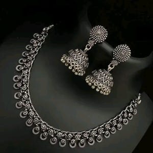 Puede incluir: Un conjunto de collar y pendientes plateados. El collar presenta una serie de pequeños colgantes circulares. Los pendientes tienen forma de campana con patrones intrincados. Las joyas se exhiben sobre una superficie negra.