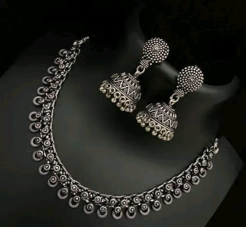 May include: A silver-toned necklace and earring set. The necklace features a series of small, circular pendants. The earrings are bell-shaped with intricate patterns. The jewelry is displayed on a black surface.