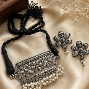 May include: A silver-toned metal necklace featuring a rectangular pendant with embossed floral and vine patterns, accented by a fringe of small white beads, suspended from a black braided cord, accompanied by matching flower-shaped stud earrings.