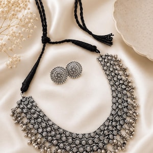 May include: A silver-toned necklace and matching stud earrings set. The necklace features a wide, curved band with intricate floral-like metalwork and small dangling silver beads along the bottom edge. It includes a black braided cord necklace strap.