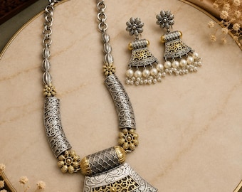 Op de afbeelding: Een sieradenset bestaande uit een ketting en bijpassende oorbellen. De stukken zijn gemaakt van geoxideerd zilver- en goudkleurig metaal met ingewikkelde filigraanpatronen, bloemaccenten en bungelende witte kunstparels en kraaltjes.