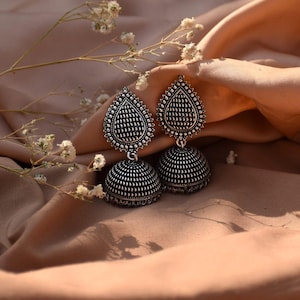 May include: A pair of silver-toned earrings with a teardrop-shaped top and a bell-shaped bottom. The earrings are decorated with small, raised dots and are displayed on a tan fabric with white flowers.