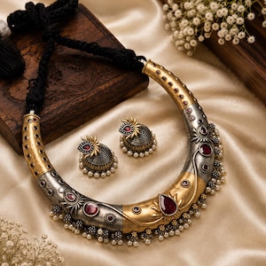 May include: A crescent-shaped necklace and matching jhumka earrings featuring a two-tone gold and oxidized silver finish. The jewelry is adorned with deep red teardrop and circular stones, intricate metal floral motifs, and small white pearl beads.