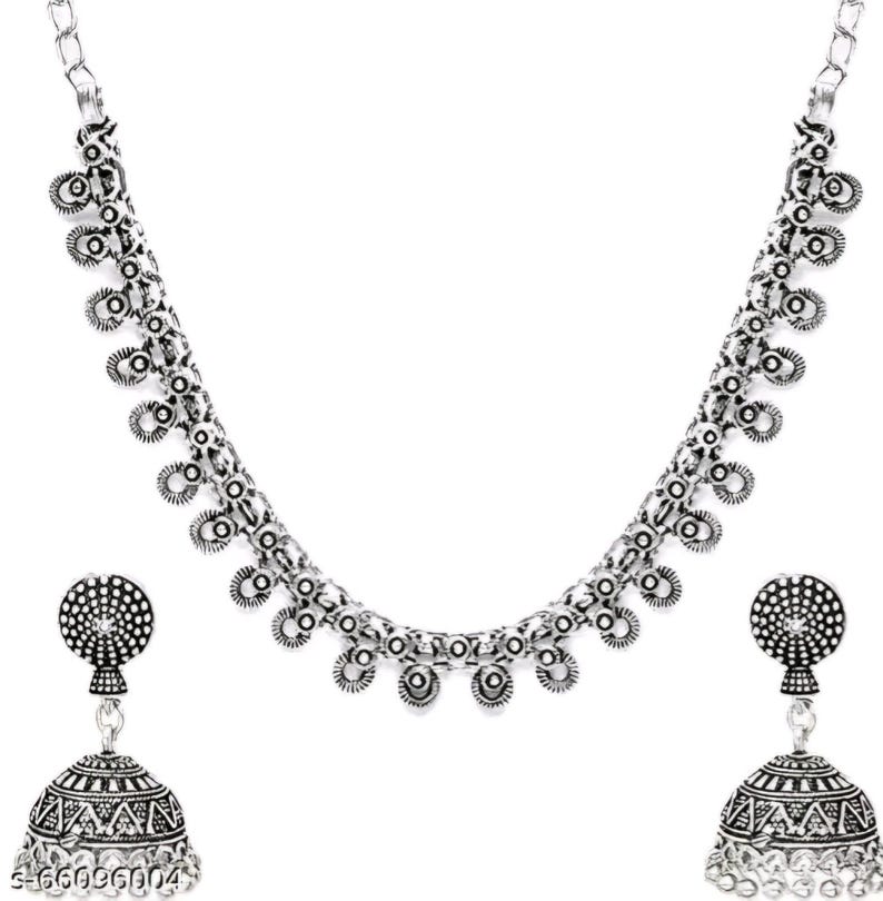 May include: A silver-toned necklace and earring set. The necklace features a chain with circular and teardrop-shaped accents. The earrings are bell-shaped with intricate patterns and a circular top. The jewelry set is displayed against a white background.