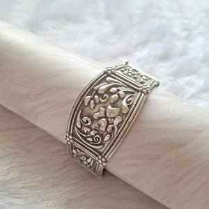 以下が含まれることがあります： 銀色のカフブレスレット。精巧な花の模様が特徴です。葉や渦巻き模様が細かく彫刻されています。金属は研磨仕上げで、白い表面に展示されています。