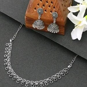 May include: A silver-toned jewelry set featuring a necklace and matching earrings. The necklace has a delicate chain with decorative elements. The earrings are bell-shaped with intricate patterns. The set is displayed on a dark surface with a wooden box and white flowers.