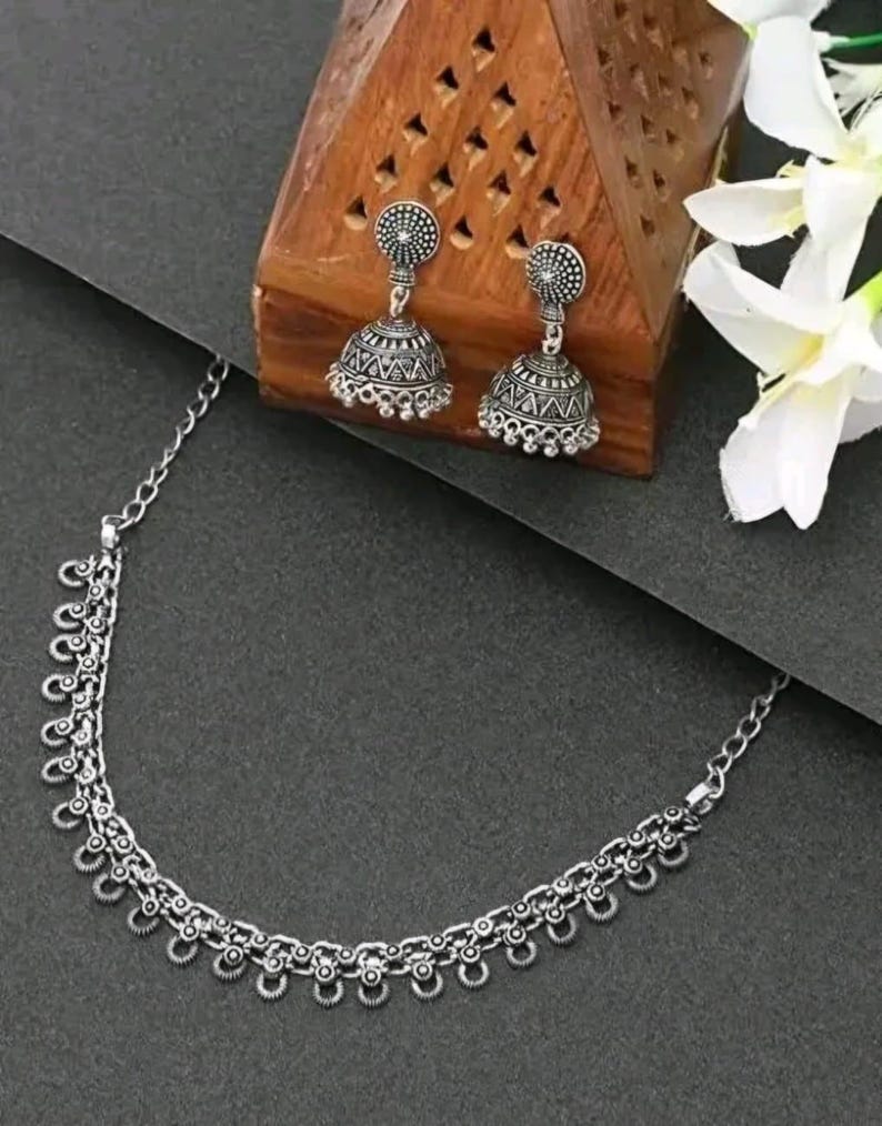 May include: A silver-toned jewelry set featuring a necklace and matching earrings. The necklace has a delicate chain with decorative elements. The earrings are bell-shaped with intricate patterns. The set is displayed on a dark surface with a wooden box and white flowers.