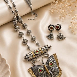 May include: A silver-toned necklace and earring set featuring a large butterfly pendant with red and green accents. The necklace includes a cylindrical centerpiece and beaded chain, while the matching earrings are small jhumka-style drops.