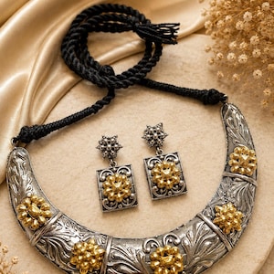 May include: A crescent-shaped silver-toned necklace with gold-toned floral accents and a black cord, accompanied by two matching rectangular drop earrings featuring a central gold-toned flower design on a silver-toned base with intricate engravings.
