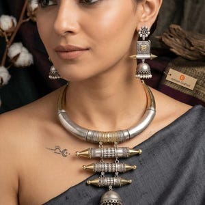 May include: A silver and gold-toned metal necklace featuring a thick curved collar, three horizontal cylindrical pendants with intricate etched patterns, and a bell-shaped jhumka drop with small white beads, paired with matching jhumka earrings.