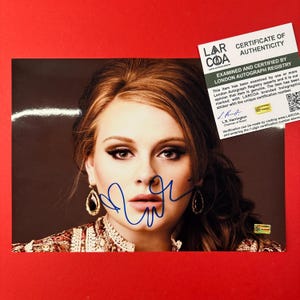 May include: A glossy photograph of Adele featuring a blue ink autograph across the center. The image includes a white LARCOA Certificate of Authenticity card in the upper right corner with black text and a small holographic sticker.