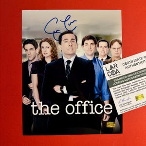 Puede incluir: Una fotografía promocional de la serie de televisión The Office con un elenco de actores, con un autógrafo en tinta azul sobre la figura central y un certificado de autenticidad blanco de London Autograph Registry a la derecha.