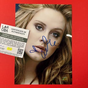 May include: A glossy photograph of a person with long blonde hair, featuring a blue ink autograph across the face. A white rectangular certificate of authenticity from LARCOA with black text and a QR code is positioned over the left side.