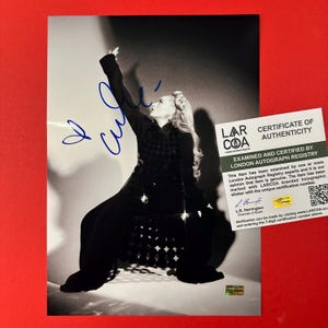 May include: A black and white photograph of a person with long hair wearing a dark outfit, posing with one arm raised. The image features a blue ink autograph across the top and a white certificate of authenticity from London Autograph Registry.