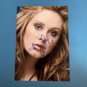 Puede incluir: Fotografía de retrato de una mujer con piel clara y cabello largo y ondulado de color marrón claro. La mujer mira directamente a la cámara. La fotografía está firmada con tinta azul. El fondo es de color azul liso.