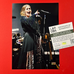 Könnte beinhalten: Ein signiertes Foto einer Person in einem schwarzen Kleid, die an einem Mikrofon auf der Bühne steht. Das Bild enthält ein weißes Echtheitszertifikat des London Autograph Registry mit schwarzem Text und QR-Code, das über dem Foto liegt.