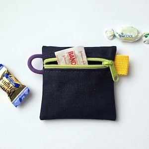 May include: A small navy blue square pouch with a lime green zip, a purple loop on the left side, and a yellow fabric tab on the right. A Band-Aid brand adhesive bandage packet is partially tucked inside the open zip compartment.