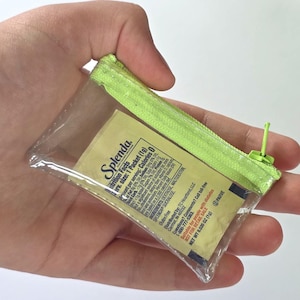 May include: A small, clear plastic pouch with a bright lime green zipper closure containing a single Splenda sweetener packet. The packet displays nutrition facts, ingredients, and distribution information printed in black text on a beige background.