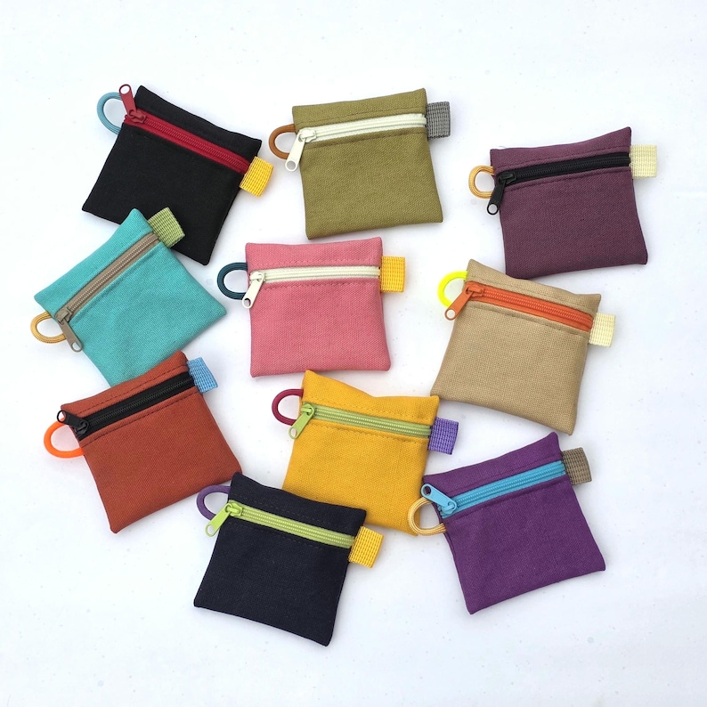 May include: A collection of eleven small, square fabric coin pouches in various solid colours including black, olive, purple, teal, pink, tan, rust, yellow, and dark blue. Each pouch features a contrasting zip closure and a small side loop.