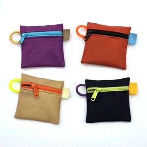 May include: Four square zipped pouches in purple, orange, tan, and black. Each pouch features a contrasting coloured zip, a matching circular pull tab on the left side, and a small fabric tab on the right side, all made of durable canvas fabric.