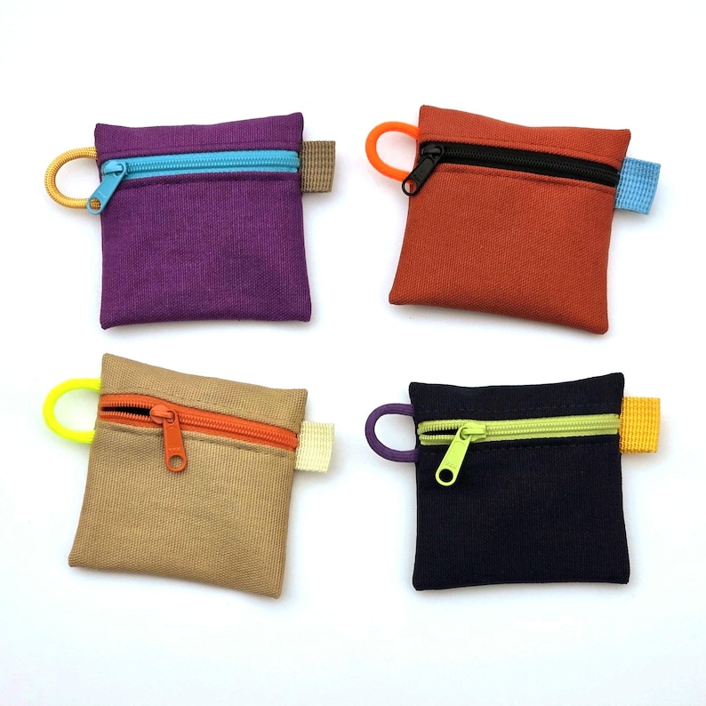 May include: Four square zipped pouches in purple, orange, tan, and black. Each pouch features a contrasting coloured zip, a matching circular pull tab on the left side, and a small fabric tab on the right side, all made of durable canvas fabric.