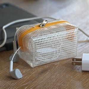 May include: A clear, mesh-like zippered pouch with an orange zipper, containing white earbuds and other small items. A white phone charger and a tablet are also visible on a wooden surface.