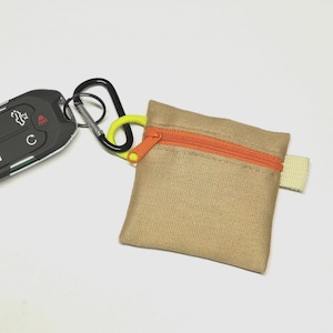 May include: A square tan fabric pouch with an orange zip, attached to a black metal carabiner and a bright yellow cord loop. A black car key fob is connected to the carabiner. The pouch features a small beige fabric tab on the right side.