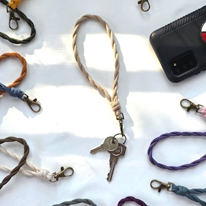 May include: A collection of braided wristlet lanyards in various solid colors including beige, yellow, dark green, brown, purple, and blue. Each lanyard features a bronze-toned metal lobster clasp and is shown with a set of silver metal keys.