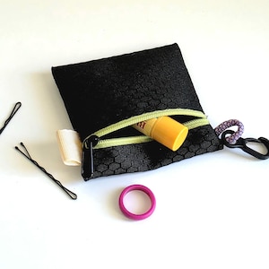 May include: Black zippered pouch with a green zipper and a small lip balm inside. The pouch has a black carabiner clip and a purple loop attached. Two black bobby pins and a pink ring are also visible.