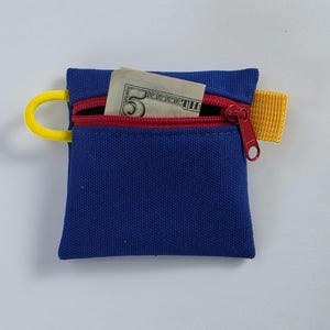 May include: A small, blue canvas pouch with a red zipper and a yellow loop and tab. A five-dollar bill is partially visible inside the open pouch. The pouch is designed to hold small items.
