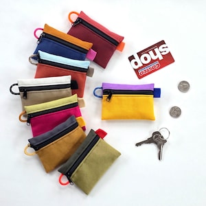 May include: A collection of small, colorful zippered pouches in various shades, including red, blue, and yellow. Each pouch has a contrasting zipper and a loop for attaching to a keychain. A Costco Wholesale card, two quarters, and a set of keys are also present.