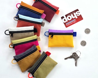 May include: A selection of small, brightly coloured zippered pouches in shades like red, blue, and yellow. Each pouch features a contrasting zip and a loop for attaching to a keyring. A Costco Wholesale card, two coins, and a set of keys are also visible.