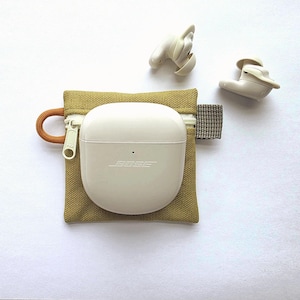 May include: A pair of off-white Bose wireless earbuds with a matching charging case resting on a small, square, olive-green canvas zipped pouch. The pouch features a light-coloured zip, an orange cord loop on the left, and a grey fabric tab on the right.