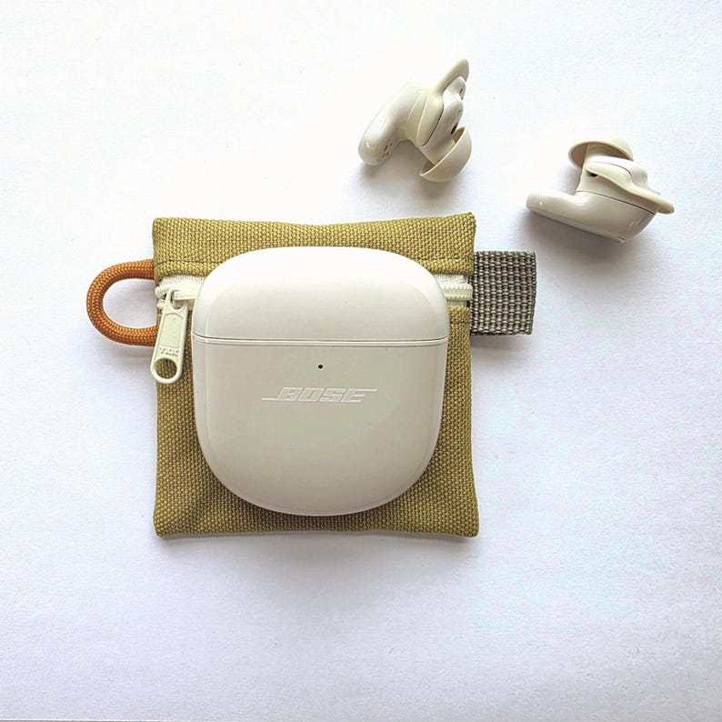 May include: A pair of off-white Bose wireless earbuds with a matching charging case resting on a small, square, olive-green canvas zipped pouch. The pouch features a light-coloured zip, an orange cord loop on the left, and a grey fabric tab on the right.